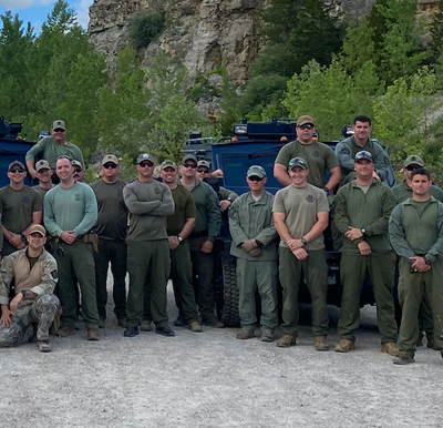A large group of tactical officers and trainees in green gear posing together outdoors in front of armored vehicles and a rocky hill.